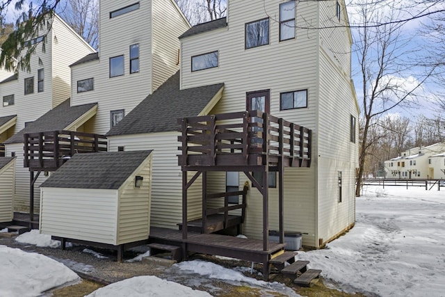 snow covered back of property with a deck, an outdoor structure, a storage unit, and roof with shingles