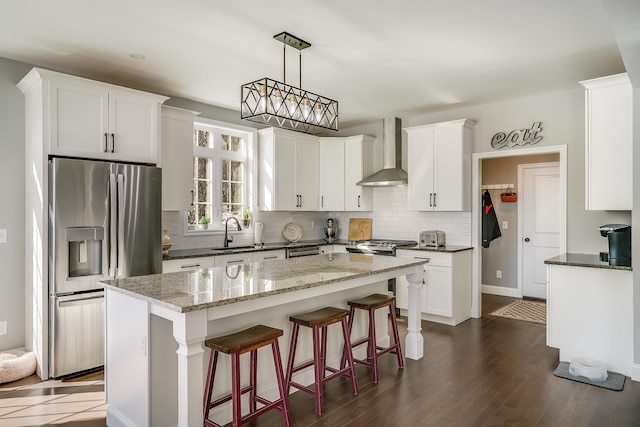 kitchen featuring a kitchen island, a sink, stainless steel appliances, white cabinetry, and wall chimney range hood