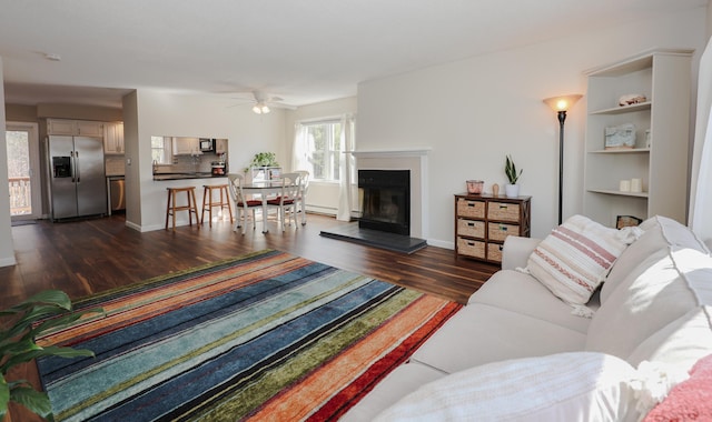 living room with baseboards, dark wood finished floors, a fireplace with raised hearth, ceiling fan, and baseboard heating