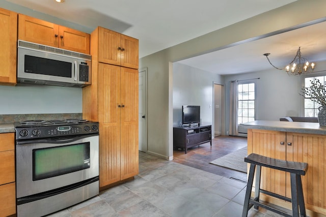 kitchen with dark countertops, a breakfast bar area, brown cabinets, appliances with stainless steel finishes, and a notable chandelier