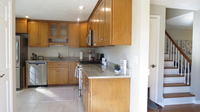 kitchen with light stone counters, a sink, stainless steel appliances, glass insert cabinets, and brown cabinets