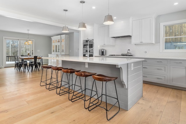 kitchen featuring white gas stovetop, premium range hood, a center island with sink, decorative backsplash, and light wood-style floors