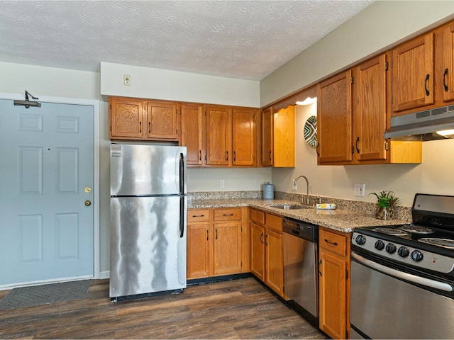 kitchen featuring brown cabinetry, a sink, dark wood-type flooring, under cabinet range hood, and appliances with stainless steel finishes