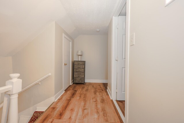 corridor with light wood-type flooring, an upstairs landing, a textured ceiling, baseboards, and vaulted ceiling