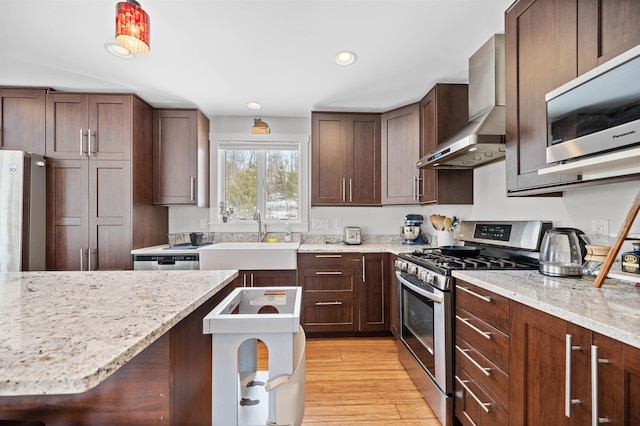 kitchen featuring light wood-style flooring, a sink, light stone counters, stainless steel appliances, and wall chimney range hood