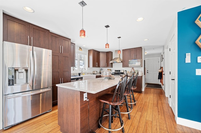 kitchen with a kitchen bar, light wood finished floors, appliances with stainless steel finishes, and wall chimney range hood