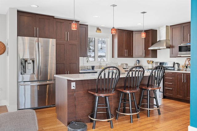 kitchen with light wood finished floors, dark brown cabinets, a breakfast bar, stainless steel appliances, and wall chimney exhaust hood