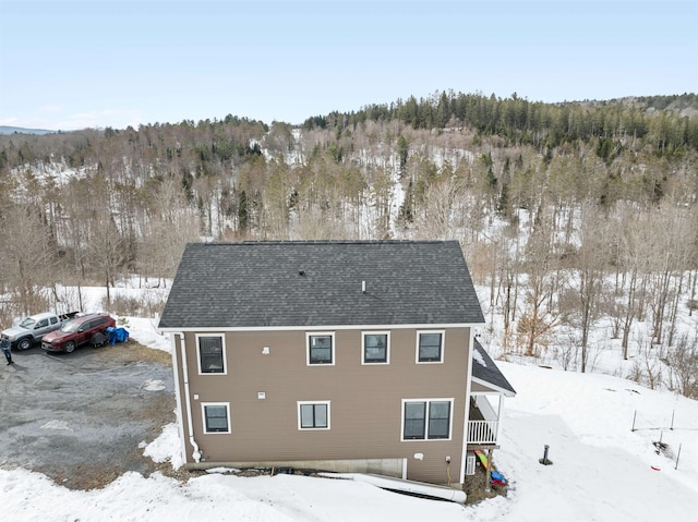 snow covered back of property featuring a forest view and a shingled roof