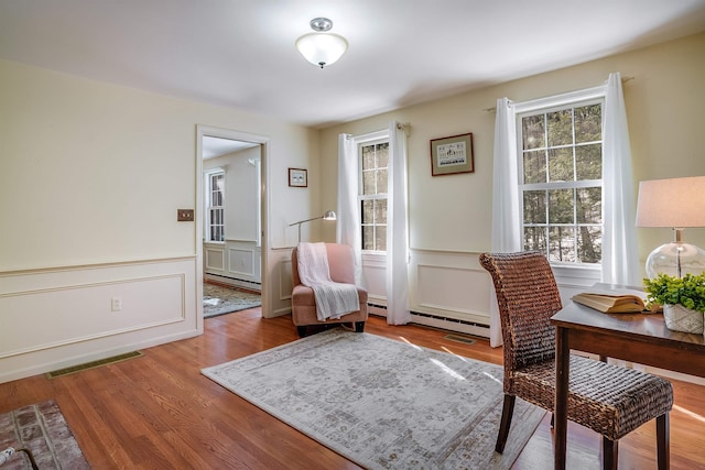 living area with a baseboard heating unit, a wainscoted wall, wood finished floors, and visible vents