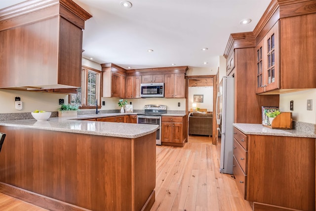 kitchen with appliances with stainless steel finishes, a peninsula, brown cabinetry, light wood finished floors, and glass insert cabinets