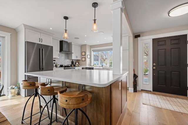 kitchen with wall chimney range hood, tasteful backsplash, freestanding refrigerator, light wood-style floors, and light countertops