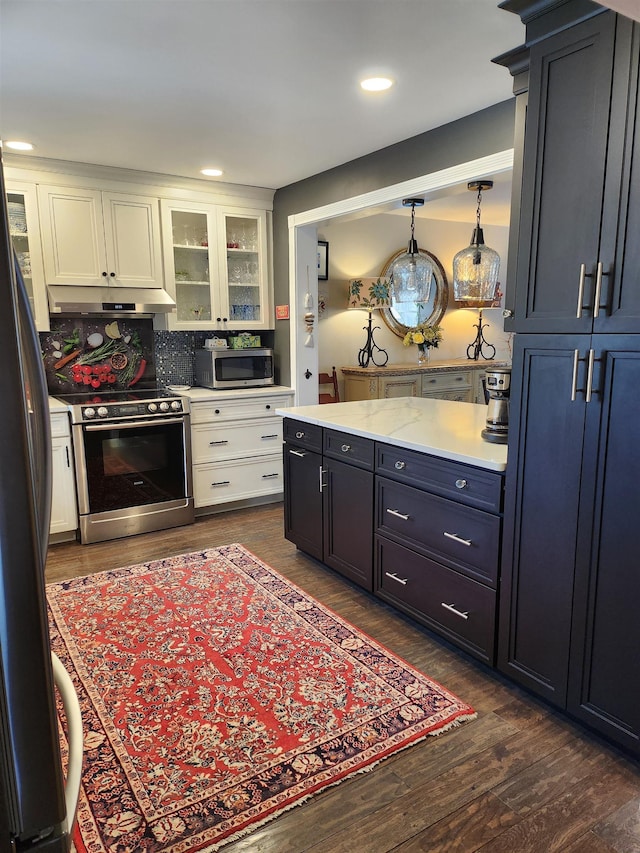 kitchen featuring dark wood-style flooring, light countertops, glass insert cabinets, under cabinet range hood, and appliances with stainless steel finishes