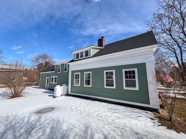 snow covered rear of property with a shingled roof and a chimney