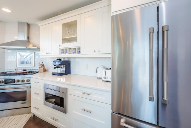 kitchen with glass insert cabinets, wall chimney range hood, light countertops, premium appliances, and decorative backsplash