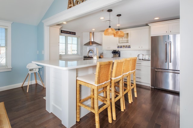 kitchen with dark wood-style floors, high end fridge, light countertops, a kitchen bar, and wall chimney range hood