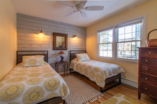 bedroom with an accent wall, wooden walls, wood finished floors, ceiling fan, and a baseboard heating unit