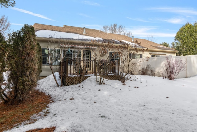 snow covered house with a deck, an attached garage, and fence