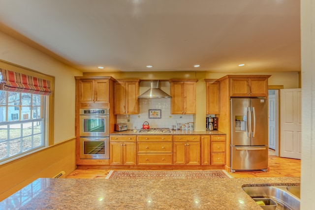 kitchen with a sink, light stone counters, backsplash, appliances with stainless steel finishes, and wall chimney exhaust hood