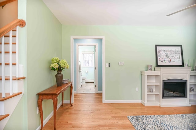 living room featuring baseboards, stairs, light wood-style floors, a glass covered fireplace, and a baseboard heating unit