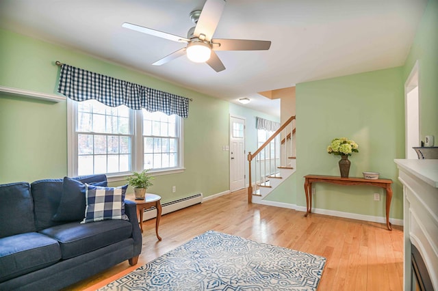 living area featuring a ceiling fan, light wood finished floors, a baseboard radiator, baseboards, and stairs