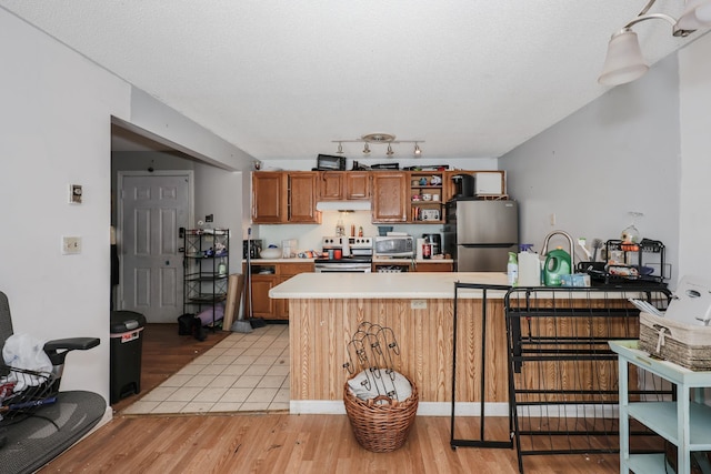 kitchen with under cabinet range hood, light countertops, appliances with stainless steel finishes, a peninsula, and brown cabinetry