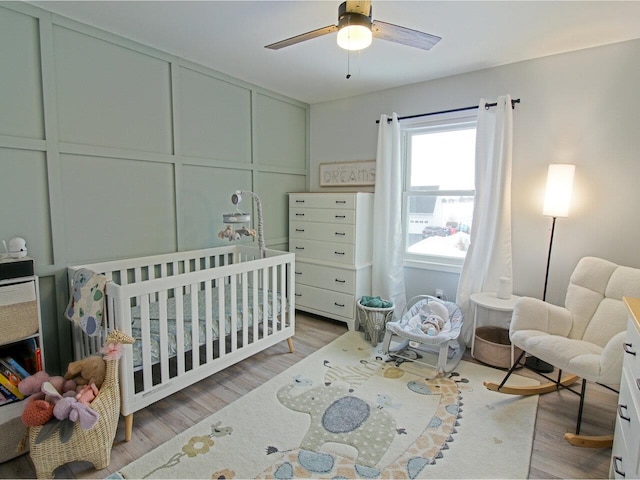 bedroom with a decorative wall, a crib, a ceiling fan, and wood finished floors