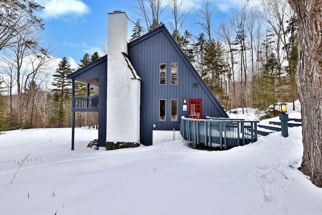 snow covered house with a wooden deck, board and batten siding, and a chimney