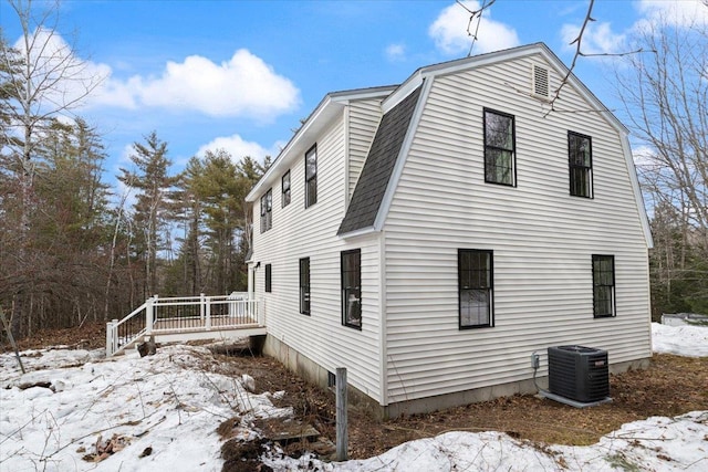 view of snow covered exterior featuring cooling unit, a gambrel roof, roof with shingles, and a wooden deck