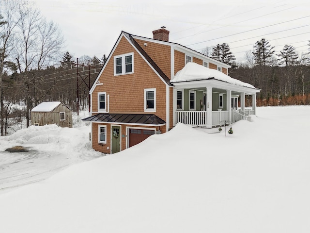 snow covered property featuring a standing seam roof, covered porch, a chimney, a garage, and metal roof