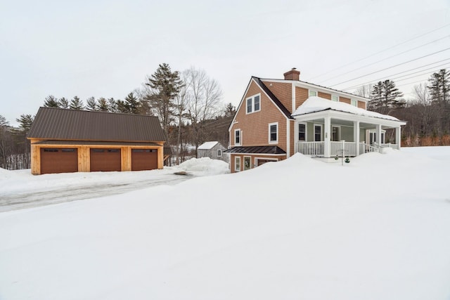 snow covered property with an outbuilding, a porch, a chimney, a garage, and metal roof