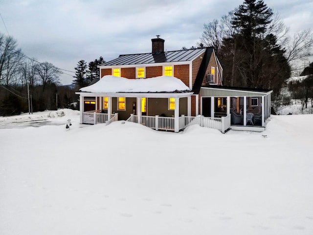 snow covered house with a porch, a chimney, metal roof, a sunroom, and a standing seam roof