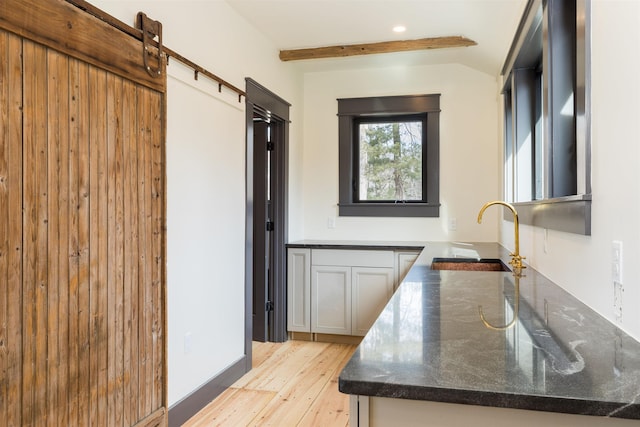 kitchen featuring light wood-type flooring, beamed ceiling, a sink, a barn door, and dark stone counters