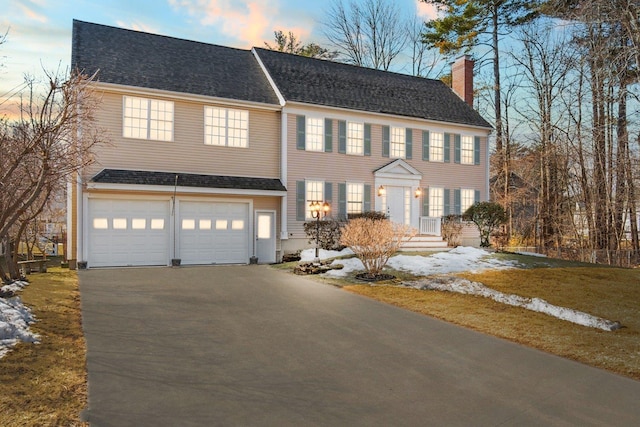 colonial-style house with a garage, roof with shingles, a chimney, and driveway