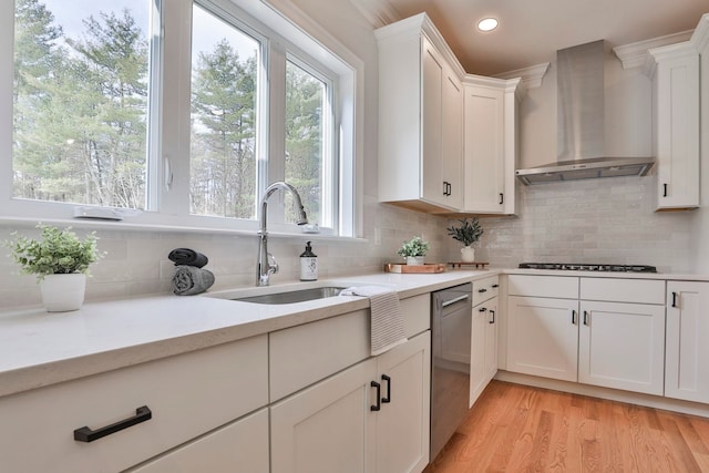 kitchen featuring wall chimney range hood, stainless steel dishwasher, white cabinets, gas cooktop, and a sink