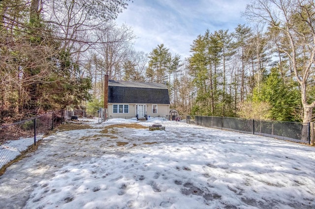 snow covered back of property featuring a chimney, a fenced backyard, a gambrel roof, and an outdoor fire pit