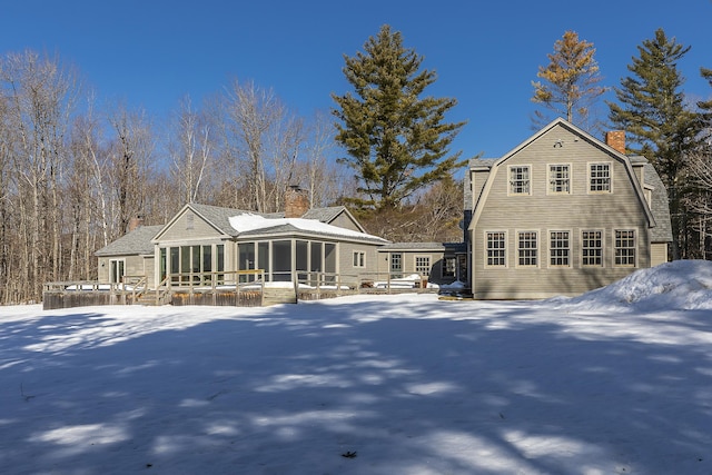 snow covered back of property with a deck, a gambrel roof, a sunroom, and a chimney