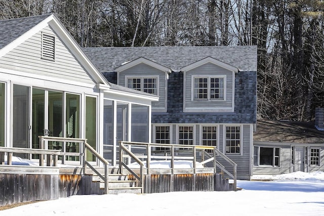 snow covered back of property with a shingled roof, a sunroom, and a wooden deck