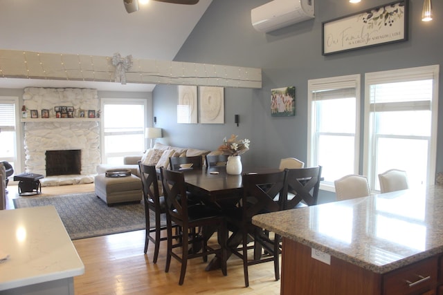dining area with light wood-style flooring, a ceiling fan, a wall mounted air conditioner, high vaulted ceiling, and a stone fireplace