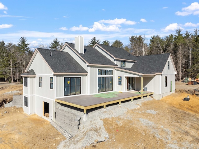 rear view of property with board and batten siding, roof with shingles, a chimney, and a deck