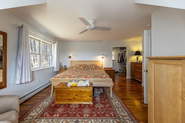 bedroom featuring wood finished floors, a ceiling fan, a baseboard heating unit, a walk in closet, and baseboards