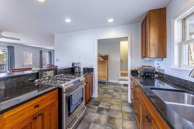 kitchen featuring a sink, dark stone counters, gas stove, stone finish flooring, and brown cabinetry