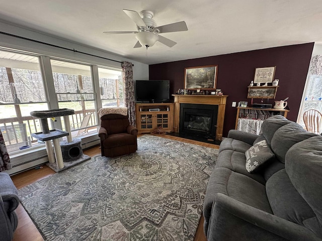 living area with a glass covered fireplace, baseboard heating, wood finished floors, and ceiling fan