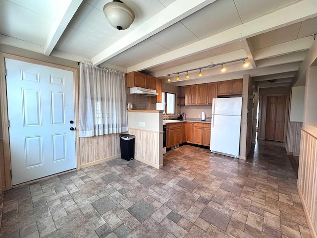 kitchen with under cabinet range hood, light countertops, wooden walls, freestanding refrigerator, and stone tile flooring