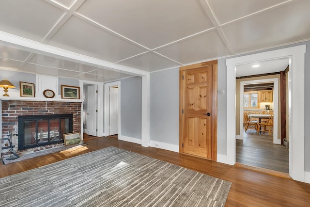 unfurnished living room with a brick fireplace, coffered ceiling, wood finished floors, and baseboards