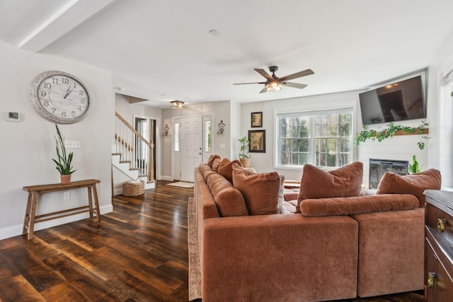 living room with a glass covered fireplace, dark wood-type flooring, stairs, a ceiling fan, and baseboards