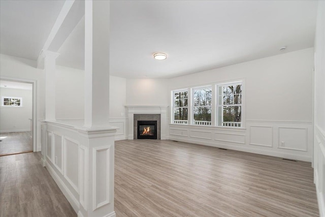 unfurnished living room with a tiled fireplace, a wainscoted wall, light wood-style flooring, a decorative wall, and ornate columns
