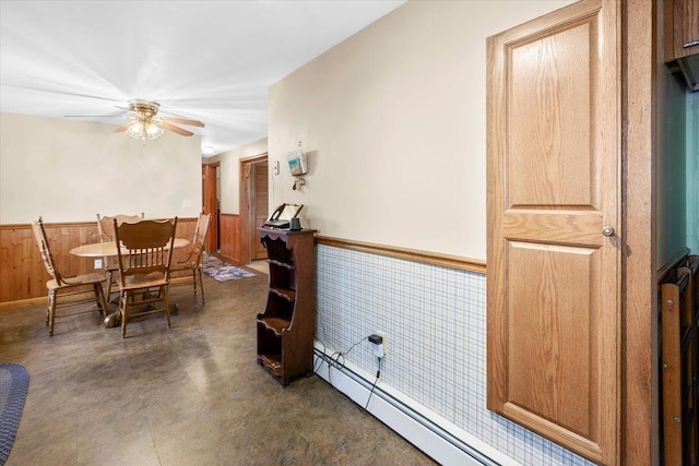 dining space with a baseboard radiator, a wainscoted wall, a ceiling fan, wooden walls, and concrete flooring