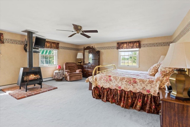 carpeted bedroom with baseboard heating, a wood stove, ceiling fan, and multiple windows