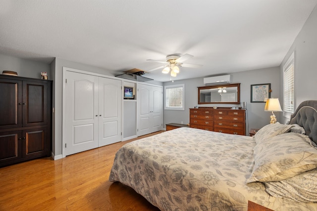 bedroom with a wall unit AC, light wood-type flooring, a ceiling fan, baseboards, and multiple closets