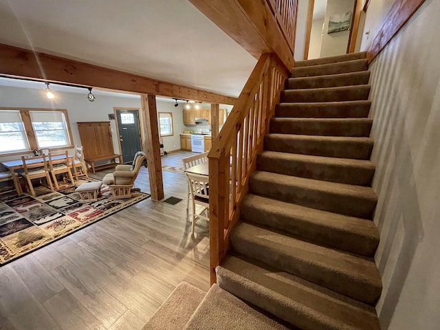stairs featuring track lighting, beamed ceiling, a wealth of natural light, and wood finished floors